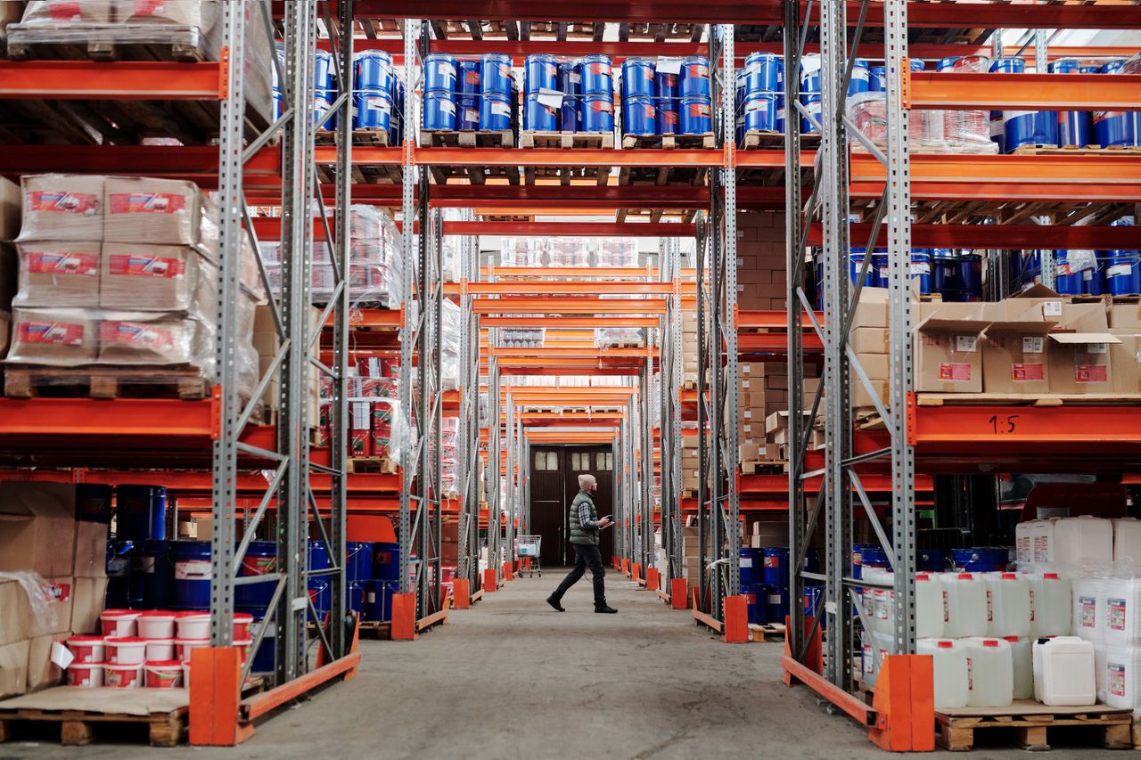 services-bg A man walking through a large industrial warehouse with stacked shelves filled with goods and products.